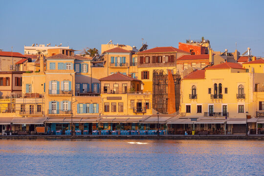 Traditional waterfront buildings and restaurants illuminated by warm sunrise light along the historic harbor of Chania, Crete, Greece.
