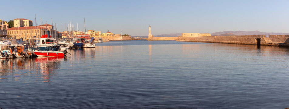 Boats and yachts in the historic harbor of Chania with the Venetian lighthouse and stone breakwater illuminated by warm sunrise light, Crete, Greece.