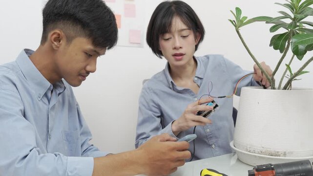 Young Asian woman and man using microcontroller and soil moisture sensors to measure soil moisture in a potted plant.