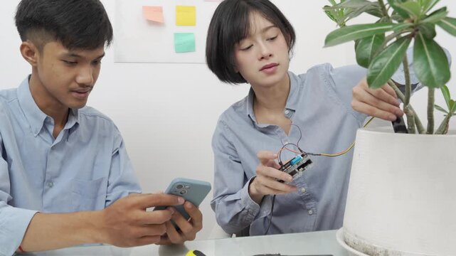 Young Asian woman and man using microcontroller and soil moisture sensors to measure soil moisture in a potted plant.