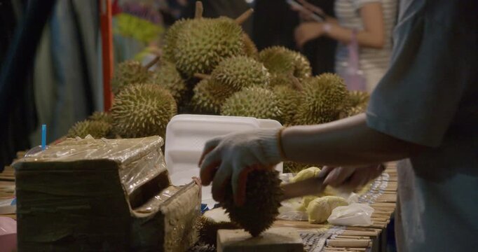 Close-up of a vendor cutting durian fruit at a Thai night market stall. Fresh tropical fruit preparation with hands, knife, and outdoor street food setting.