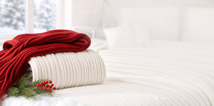Folded red and white knitted blankets on bed with pine sprig. Cozy winter bedroom interior with snowy window view