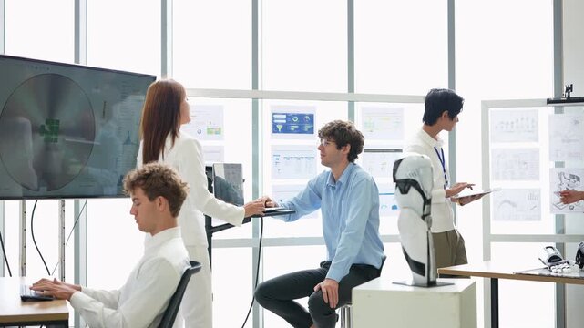 Engineer in blue shirt on a stool turns to explain to a colleague with a tablet while a woman in white suit studies the wafer display beside him, robotics R and D