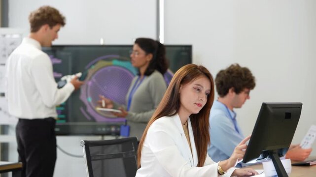 Woman in white blazer rests her chin on her hand at her desk while a young engineer holding a bionic hand talks with an Indian colleague at a circular scan screen