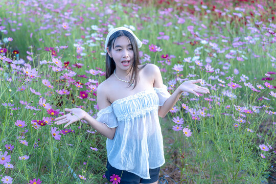 Beautiful young Asian woman in a white top posing amidst a vibrant pink and purple cosmos flower field in nature, wearing a headscarf during sunny outdoor leisure travel vacation in a scenic landscape