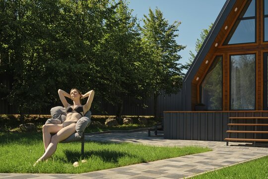 Young woman in a bathing suit sunbathing while sitting in a soft chair on the lawn at backyard of tiny house