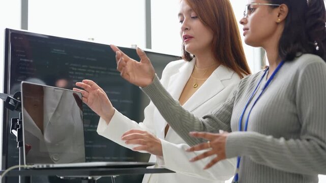 Woman in white blazer explains an idea at a laptop stand while an Indian female colleague with glasses listens and gestures, during an iterative design check, an embodied AI research scene