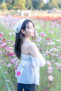 Beautiful young Asian woman in a white top posing amidst a vibrant pink and purple cosmos flower field in nature, wearing a headscarf during sunny outdoor leisure travel vacation in a scenic landscape