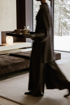 A young caucasian woman in flowing dark clothing carries a tea tray through a modern winter retreat overlooking snowy woods. Motion blur, selective focus.