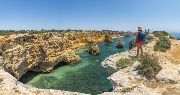 Young woman standing on rocks at steep coast, view over turquoise sea, beach Praia da Marinha, jagged rocky coast of sandstone, rock formations in the sea, Algarve, Lagos, Portugal