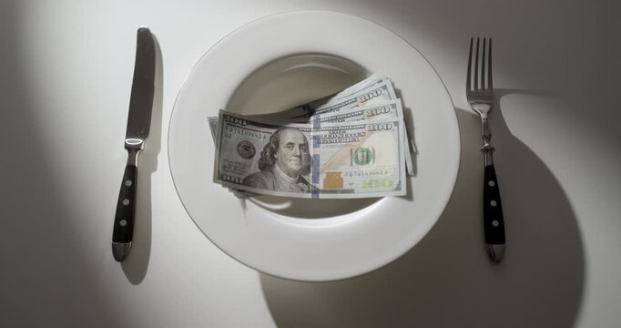 White plate filled with 100 US dollar banknotes arranged like a meal on a white table with a fork and knife. Creative metaphor for extreme wealth, luxury, financial success, greed, money obsession.