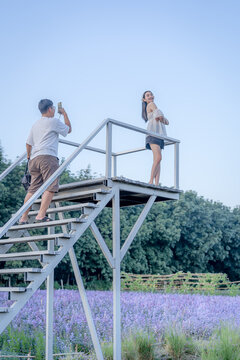 Full body portrait of a young Asian boy taking a photo of a young woman posing on a white metal observation tower platform overlooking a scenic lavender field during a clear day with a bright blue sky