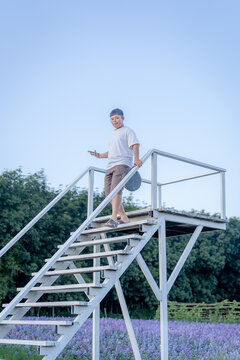 Full body portrait of a young Asian boy walking down a white metal outdoor staircase holding a smartphone in a scenic lavender field during a clear day with a bright blue sky background