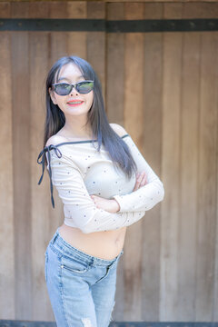 Portrait of a trendy young Asian woman wearing stylish black sunglasses and a polka dot top, crossing her arms and smiling against a rustic wooden wall background during a sunny leisure day