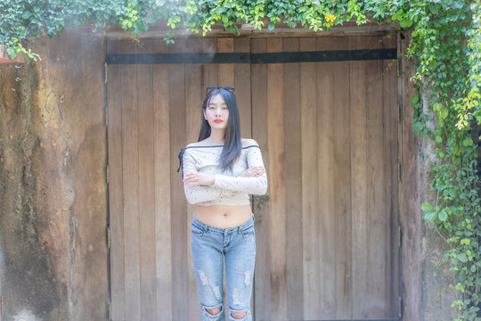 Full body portrait of a trendy young Asian woman crossing her arms and smiling against a rustic wooden door background framed with lush green ivy leaves during a sunny leisure day in an outdoor settin