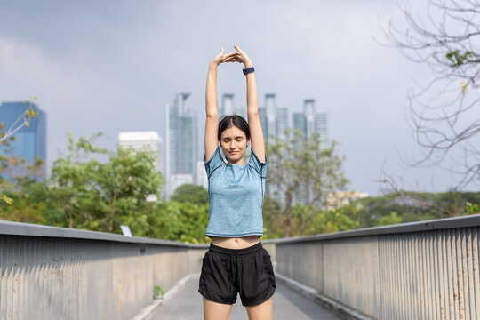 Woman stretching arms overhead on urban bridge before outdoor exercise, city skyline background, warm up, flexibility training, sportswear, smartwatch, mobility, wellness and healthy routine.