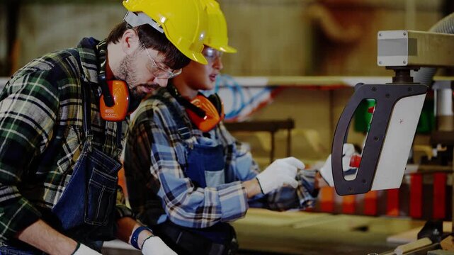 Bearded carpenter and curly haired female coworker in yellow hard hats measure a plywood panel together next to a sliding table saw inside a timber and plywood factory