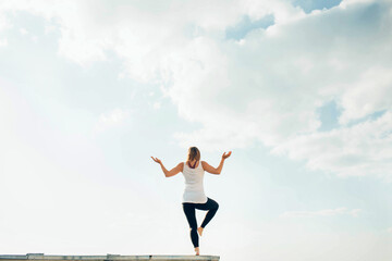 Obraz premium Young woman practices yoga outside. Blonde girl turned back standing on one leg, second raised bent at knee. Hands on sides bent at elbows and turned up. Sky and clouds on background.