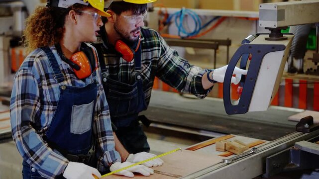 Bearded supervisor and a female trainee bend over the saw table together, lining up a board and checking the cut mark with a tape measure, in a timber factory, 4K