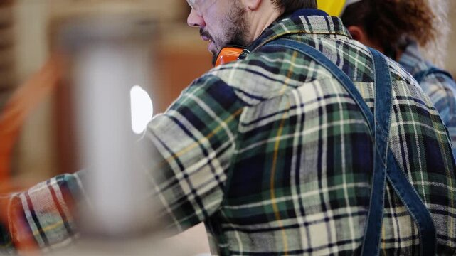 Over the shoulder shot behind a bearded carpenter in plaid and denim bib overalls as he works beside a female colleague at the saw inside a timber and plywood factory