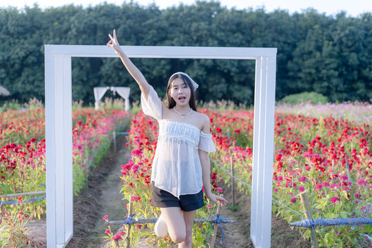 Beautiful young Asian woman posing with a victory sign inside a white archway in a colorful blooming flower garden with red and yellow blossoms during sunny outdoor leisure travel vacation in nature