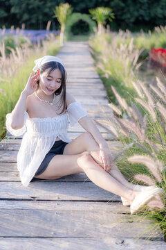 Portrait of a trendy young Asian woman sitting on a wooden pathway in a scenic outdoor garden with blooming flowers during a sunny day, wearing a white top and a headscarf with a gentle smile