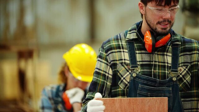 Bearded carpenter in safety glasses measures the edge of a plywood board with a yellow tape as his female colleague rests her chin on her hand behind, real time