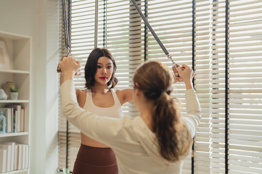 Pilates instructor and her client reach overhead together, both pulling resistance handles in perfect sync. This close-up moment captures shared focus, guided technique, and the connection between coa