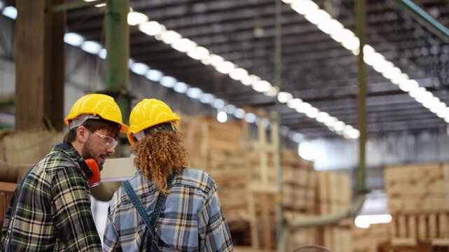 Rear view of a bearded man and a curly haired woman in yellow hard hats walking to a machine under long rows of factory ceiling lights, real time 4K footage of timber industry work