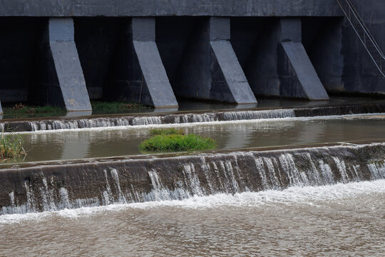 Massive grey concrete dam spillway structure with water cascading over tiered stone barriers and small green vegetation island in river bed
