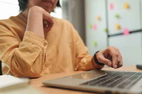 Thoughtful Asian female freelancer working on a laptop while considering a problem at home. Focused decision making and productivity.