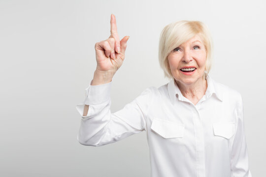 Nice picture of old woman standing at the white wall and pointing up. A brilliant idea has come up to her mind. Now she wants to materialize it. Isolated on white background