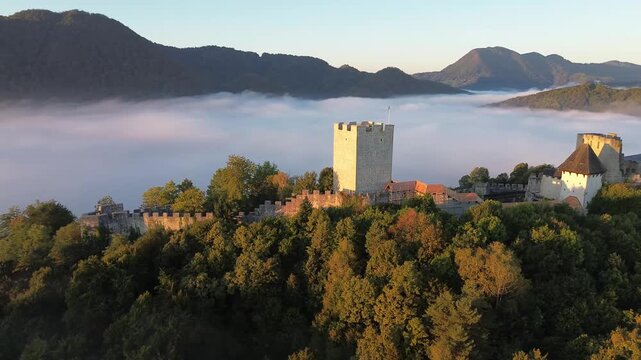 Aerial view of Celje Castle in Slovenia, surrounded by misty forested hills and mountains, creating a dramatic medieval landscape.
