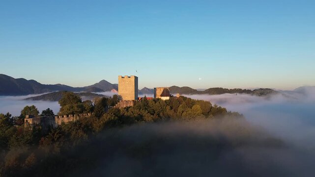 Aerial drone view of Celje Castle surrounded by autumn trees and rising above a layer of fog, with mountains in the background in Slovenia.