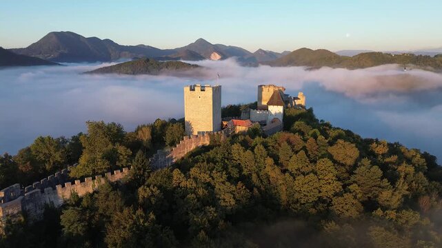 Aerial view of Celje Castle rising above a sea of fog in Slovenia, with medieval stone walls, autumn trees and dramatic mountain peaks illuminated by warm morning light.
