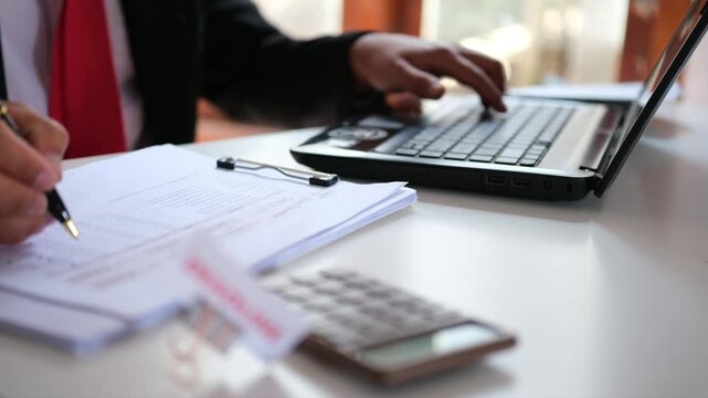 An accountant appears busy at their desk, scrolling through a laptop while cross-referencing the screen with documents during an audit.
