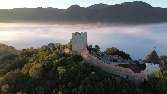Aerial view of Celje Castle rising above a sea of fog in Slovenia, with medieval stone walls, autumn trees and dramatic mountain peaks illuminated by warm morning light.