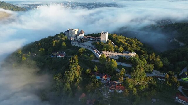 Static aerial view of Celje Castle surrounded by golden autumn forest and drifting fog, with medieval fortress walls and red-roofed houses visible below in Slovenia.

