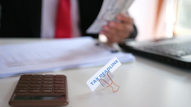 The text "TAX DEADLINE" set against a background of an office worker calculating figures on a calculator. Concept of tax payment deadlines.
