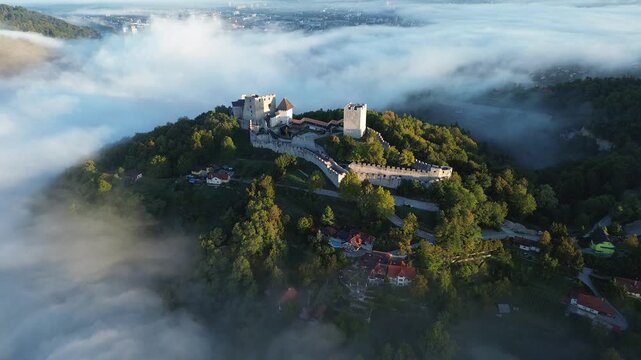 Aerial drone view of Celje Castle surrounded by a sea of fog, with medieval fortress walls, a forested hill and red-roofed houses visible below in Slovenia.
