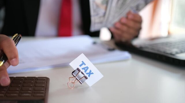 The word "TAX" on a sheet of paper clipped to a desk. Tax season concept, featuring an office worker calculating a budget with a calculator in the background. Concept of tax payment planning and calcu