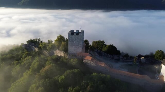 Aerial view of Celje Castle surrounded by fog in Slovenia, with medieval towers and walls emerging above the mist, autumn trees and mountain peaks in the background.
