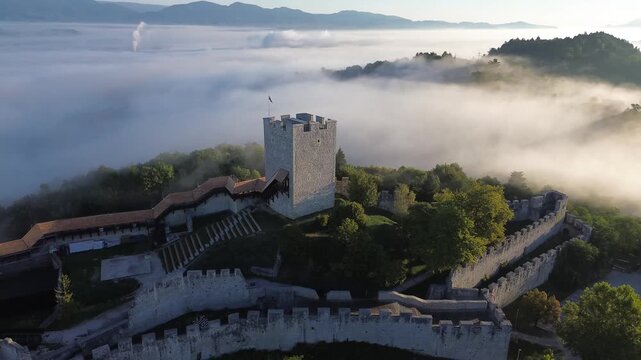 Aerial drone view of Celje Castle and Frederick's Tower above a layer of fog, with medieval fortress walls, forested hills and distant mountains in Slovenia.
