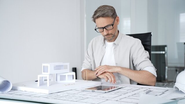 Male architect working on architectural plans and a house model, using a digital tablet for design and planning in a bright contemporary office environment