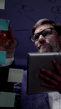 Mature male professional, wearing eyeglasses and suit, working late in a dark office, holding a digital tablet and brainstorming new business strategies with post it notes on a glass board
