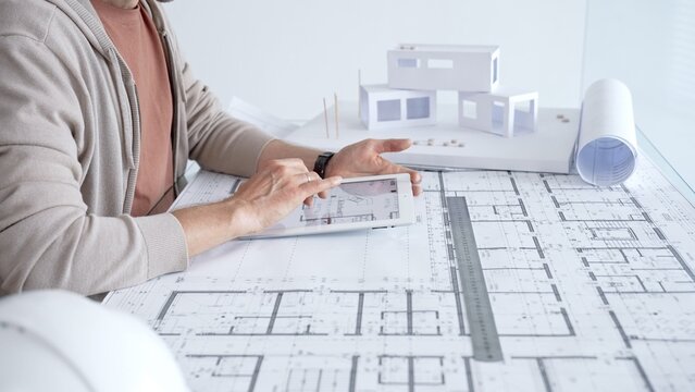Architect working on a new building design using a tablet with digital blueprint plans. Also showing paper blueprints. A ruler. And a scale model of a house on the desk