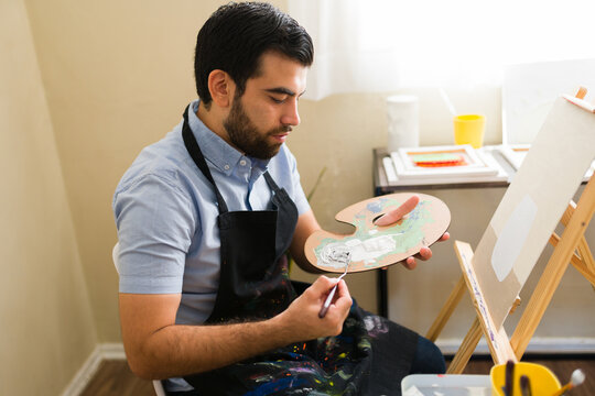 Latin man painting on canvas during an art class, mixing colors on palette with a brush, enjoying creative hobby