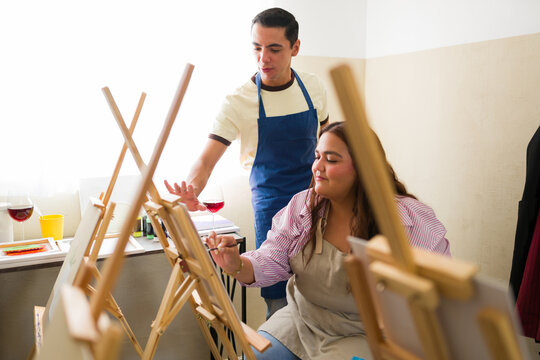Plus size woman painting on easel, receiving instruction from male teacher in an art workshop, drinking red wine