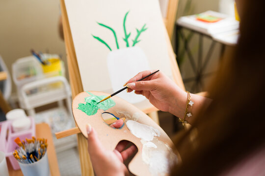 Woman learning painting in adult art class, holding palette and brush, mixing green paint on canvas