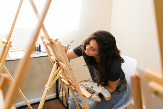 Young woman learning to paint in an adult art class, holding a palette and brush, concentrating on her canvas